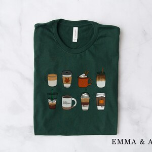 Może przedstawiać: Zielony T-shirt z nadrukiem ośmiu r&oacute;żnych napoj&oacute;w kawowych, w tym latte z przyprawami dyniowymi, caramel macchiato i latte z liściem klonowym. Tekst "Emma & Anna" jest nadrukowany w prawym dolnym rogu koszulki.