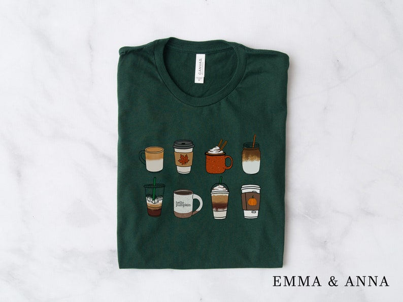 Może przedstawiać: Zielony T-shirt z nadrukiem ośmiu r&oacute;żnych napoj&oacute;w kawowych, w tym latte z przyprawami dyniowymi, caramel macchiato i latte z liściem klonowym. Tekst "Emma & Anna" jest nadrukowany w prawym dolnym rogu koszulki.