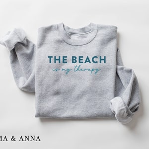 以下が含まれることがあります： 青い文字で「THE BEACH is my therapy」と書かれた灰色のスウェットシャツ