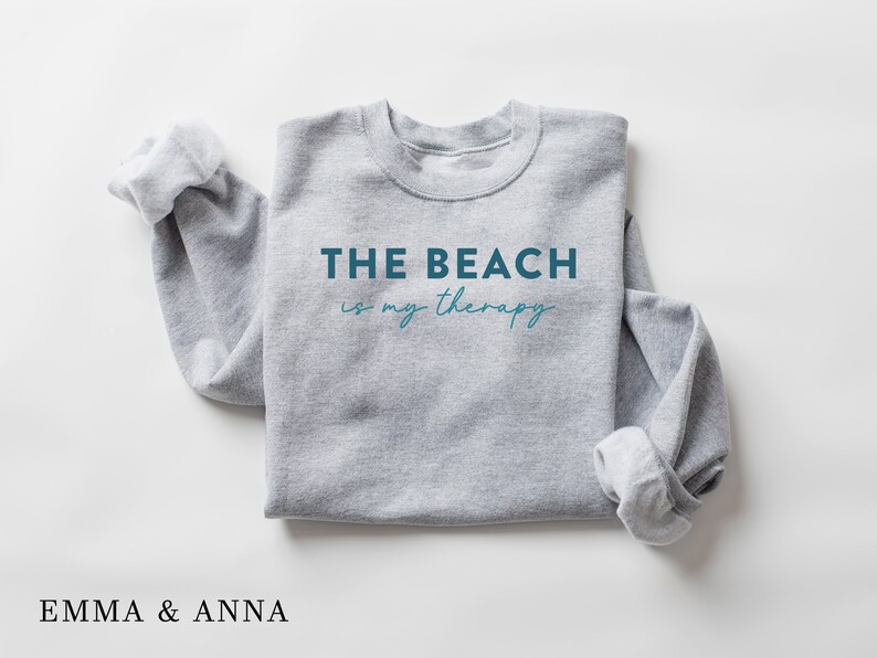 以下が含まれることがあります： 青い文字で「THE BEACH is my therapy」と書かれた灰色のスウェットシャツ