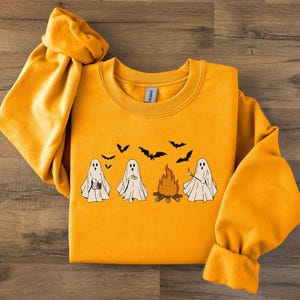 May include: Mustard yellow sweatshirt with a Halloween graphic. The design shows four cartoon ghosts, a campfire, and bats. The ghosts are holding different items. The sweatshirt has long sleeves and a crew neck.