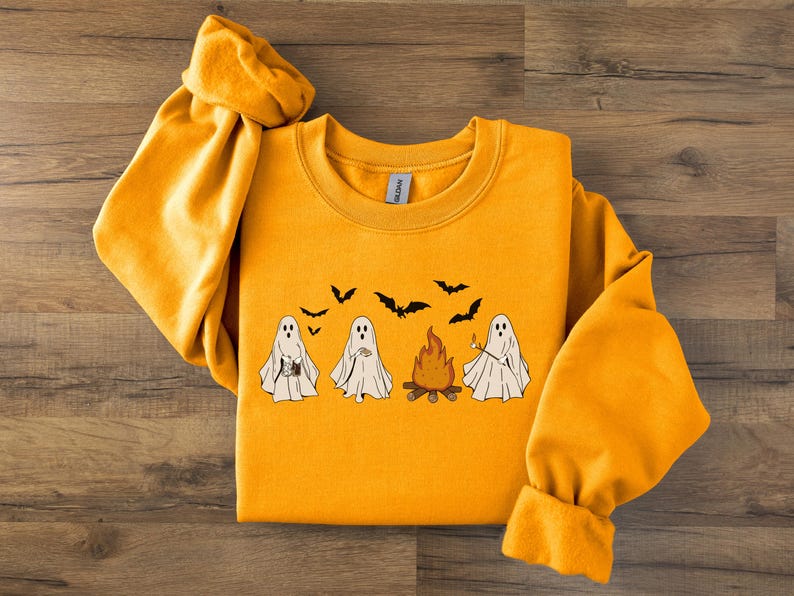 May include: Mustard yellow sweatshirt with a Halloween graphic. The design shows four cartoon ghosts, a campfire, and bats. The ghosts are holding different items. The sweatshirt has long sleeves and a crew neck.