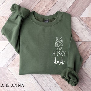 Pode incluir: Uma camisa de moletom verde com um contorno branco de um c&atilde;o husky e o texto "HUSKY dad" na frente.