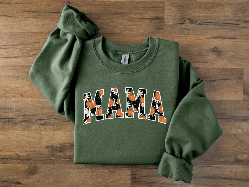 Op de afbeelding: Een olijfgroene sweatshirt met het woord "MAMA" in een witte, gebogen letter. De letters zijn gevuld met een patroon van oranje pompoenen en witte madeliefjes op een zwarte achtergrond. De sweatshirt heeft lange mouwen.
