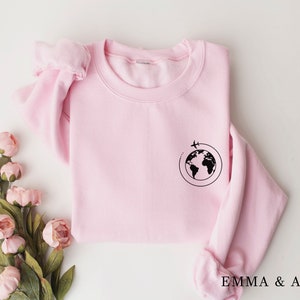 May include: A pink sweatshirt with a black graphic of a globe with a plane flying around it. The text "EMMA & ANNA" is printed on the bottom of the sweatshirt.
