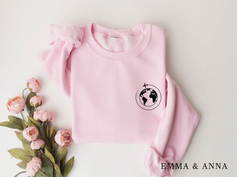 May include: A pink sweatshirt with a black graphic of a globe with a plane flying around it. The text "EMMA & ANNA" is printed on the bottom of the sweatshirt.