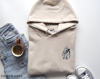 Op de afbeelding: Een beige hoodie met een zwarte geest die een kleine zwarte kat vasthoudt op de voorkant. De hoodie ligt op een wit oppervlak met een blauwe spijkerbroek en een bruine koffiemok.