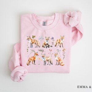May include: Pink sweatshirt with a watercolor design of foxes and flowers. The foxes are orange and white with black noses and eyes. The flowers are a variety of colors, including pink, yellow, and blue.