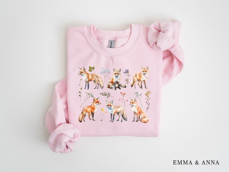 May include: Pink sweatshirt with a watercolor design of foxes and flowers. The foxes are orange and white with black noses and eyes. The flowers are a variety of colors, including pink, yellow, and blue.