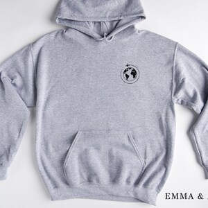 May include: A gray hooded sweatshirt with a small black graphic of a globe with a plane flying over it. The text "EMMA & ANNA" is printed on the bottom right corner of the image.