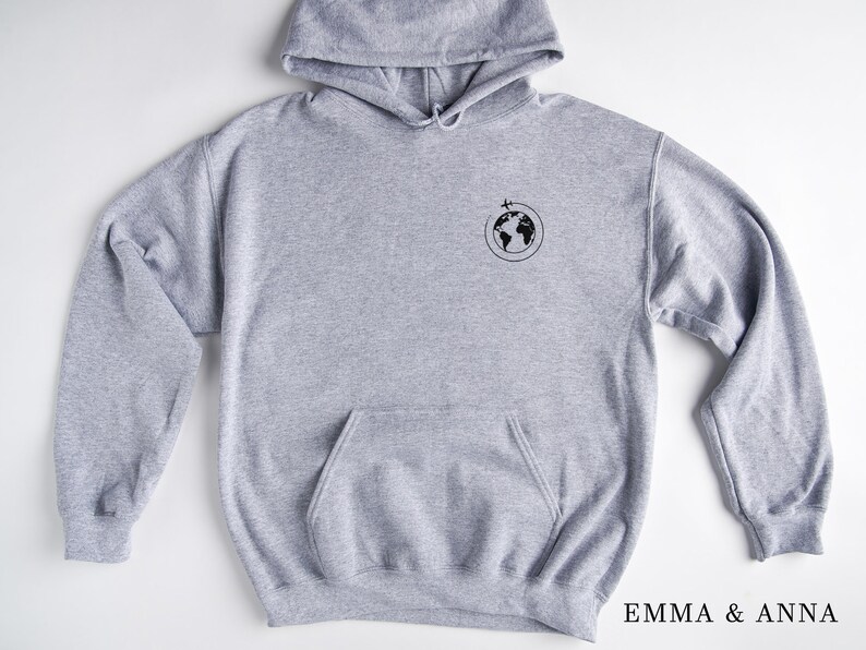 May include: A gray hooded sweatshirt with a small black graphic of a globe with a plane flying over it. The text "EMMA & ANNA" is printed on the bottom right corner of the image.