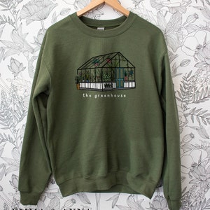 May include: An olive green sweatshirt with a greenhouse illustration. The design shows a greenhouse with a white picket fence, a blue door, and the text "the greenhouse". The greenhouse is filled with plants.