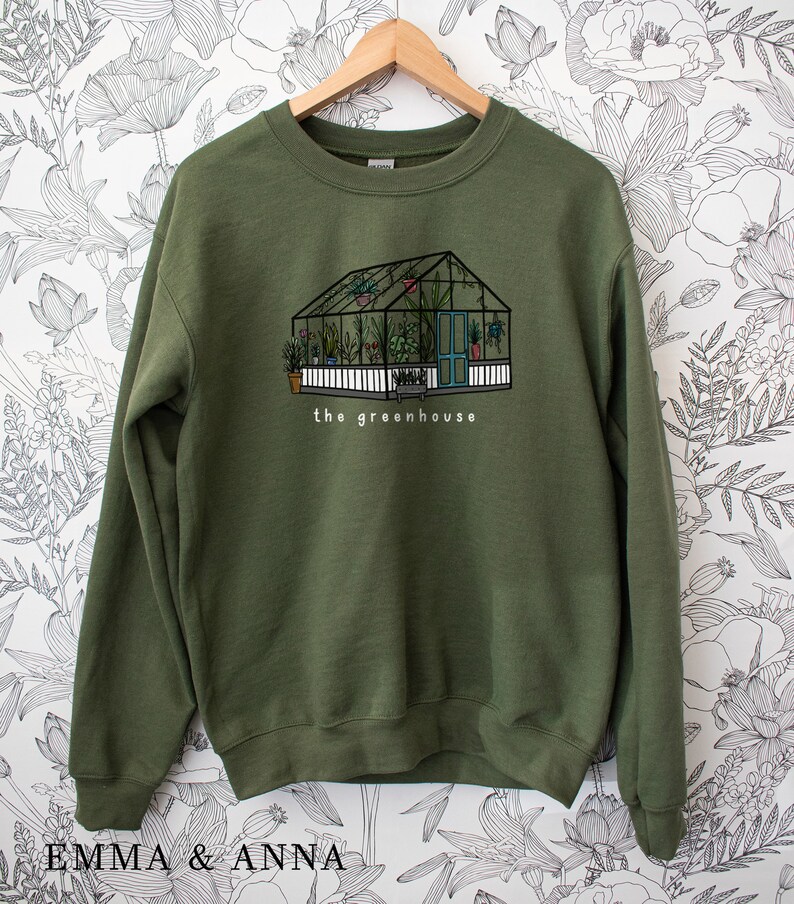 May include: An olive green sweatshirt with a greenhouse illustration. The design shows a greenhouse with a white picket fence, a blue door, and the text "the greenhouse". The greenhouse is filled with plants.