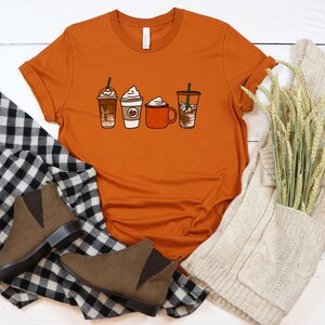 Puede incluir: Una camiseta naranja quemada con un gr&aacute;fico de cuatro bebidas de caf&eacute; diferentes. Las bebidas incluyen un caramel macchiato, un latte de especias de calabaza, un chocolate caliente y un caf&eacute; helado. La camiseta est&aacute; colocada sobre una superficie de madera blanca con una bufanda de punto blanca y una bufanda de cuadros blancos y negros.