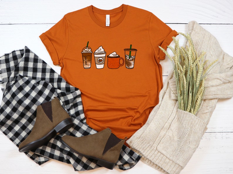 Puede incluir: Una camiseta naranja quemada con un gr&aacute;fico de cuatro bebidas de caf&eacute; diferentes. Las bebidas incluyen un caramel macchiato, un latte de especias de calabaza, un chocolate caliente y un caf&eacute; helado. La camiseta est&aacute; colocada sobre una superficie de madera blanca con una bufanda de punto blanca y una bufanda de cuadros blancos y negros.