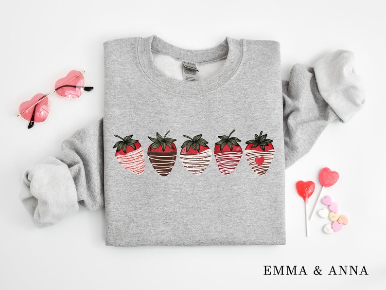 Puede incluir: Sudadera gris con un gr&aacute;fico de cinco fresas cubiertas de chocolate con un chorrito blanco. Las fresas est&aacute;n dispuestas en una fila a trav&eacute;s del centro de la sudadera.