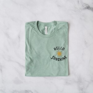 May include: A folded sage green t-shirt with the text "HELLO SUNSHINE" and a yellow sun graphic.