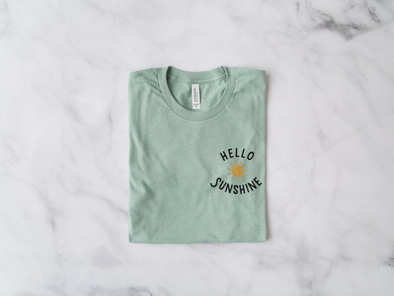 May include: A folded sage green t-shirt with the text "HELLO SUNSHINE" and a yellow sun graphic.