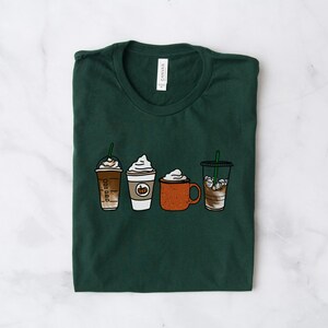 Puede incluir: Una camiseta verde oscuro con un gr&aacute;fico de cuatro bebidas de caf&eacute; diferentes. Las bebidas incluyen un latte de especias de calabaza, un caramel macchiato, un chocolate caliente y un caf&eacute; helado.