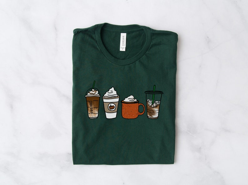 Puede incluir: Una camiseta verde oscuro con un gr&aacute;fico de cuatro bebidas de caf&eacute; diferentes. Las bebidas incluyen un latte de especias de calabaza, un caramel macchiato, un chocolate caliente y un caf&eacute; helado.