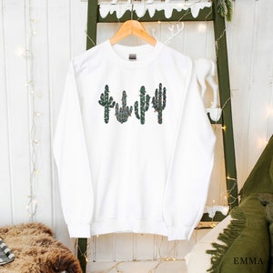 May include: White sweatshirt with a green, red, and white cactus design. The cactus is decorated with small red and white flowers.