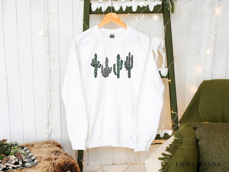 May include: White sweatshirt with a green, red, and white cactus design. The cactus is decorated with small red and white flowers.
