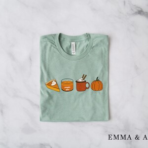 May include: A light green t-shirt with a graphic of a pumpkin, a candle that says "pumpkin spice", a mug of hot chocolate with whipped cream, and a slice of pumpkin pie.