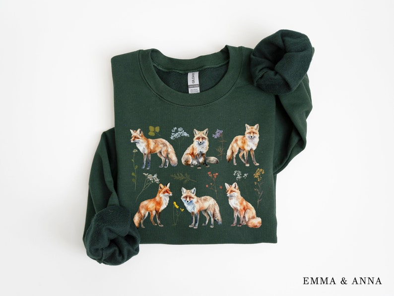 May include: A dark green sweatshirt with a repeating pattern of foxes and wildflowers. The foxes are orange and white with black noses and eyes. The wildflowers are in shades of pink, purple, yellow, and green.