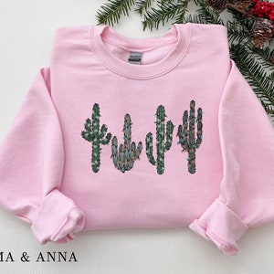 May include: Pink sweatshirt with a graphic of four cacti with Christmas lights on them.