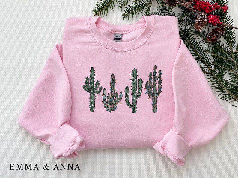 May include: Pink sweatshirt with a graphic of four cacti with Christmas lights on them.