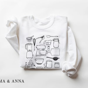 May include: White sweatshirt with a black and white illustration of kitchen tools and appliances, including a blender, mixer, measuring cups, spoons, and a whisk. The text "EMMA & ANNA" is printed below the illustration.