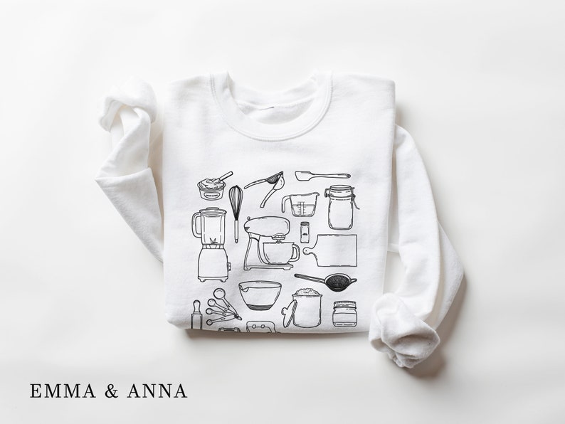 May include: White sweatshirt with a black and white illustration of kitchen tools and appliances, including a blender, mixer, measuring cups, spoons, and a whisk. The text "EMMA & ANNA" is printed below the illustration.