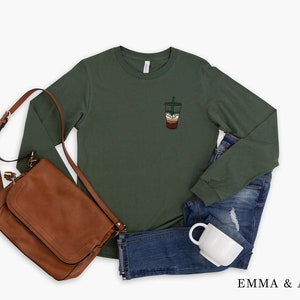 Peut inclure: Un t-shirt vert &agrave; manches longues avec une petite image brod&eacute;e d'un gobelet &agrave; caf&eacute; avec une paille verte. Le t-shirt est pr&eacute;sent&eacute; sur un fond blanc avec un sac en cuir marron, un jean bleu et une tasse blanche.