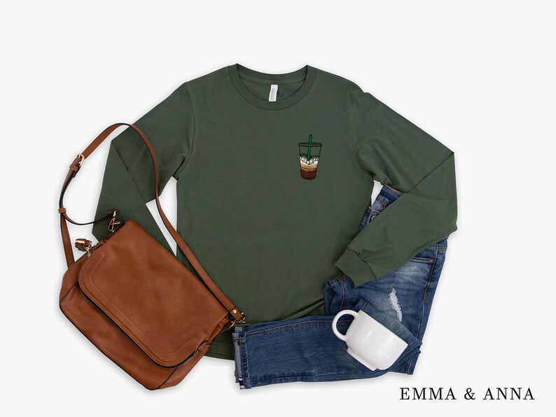 Peut inclure: Un t-shirt vert &agrave; manches longues avec une petite image brod&eacute;e d'un gobelet &agrave; caf&eacute; avec une paille verte. Le t-shirt est pr&eacute;sent&eacute; sur un fond blanc avec un sac en cuir marron, un jean bleu et une tasse blanche.