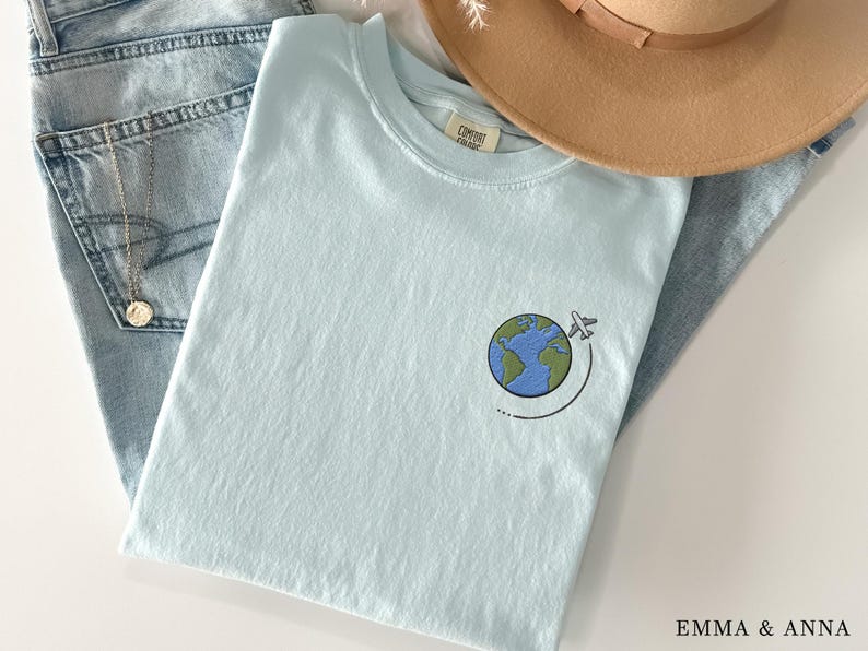 May include: A light blue t-shirt with an embroidered design of a globe with a plane flying around it. The text "Comfort Colors" is visible on the tag.