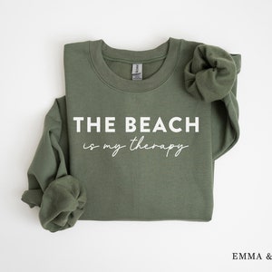 以下が含まれることがあります： 白い文字で「THE BEACH is my therapy」と書かれた緑色のスウェットシャツ。スウェットシャツは折り畳まれ、袖はまくり上げられています。