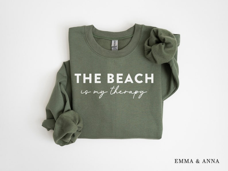 以下が含まれることがあります： 白い文字で「THE BEACH is my therapy」と書かれた緑色のスウェットシャツ。スウェットシャツは折り畳まれ、袖はまくり上げられています。