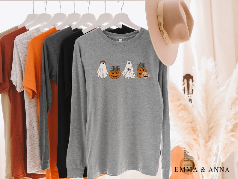 May include: A gray long-sleeve shirt with a graphic of three ghosts holding pumpkins. The ghosts are wearing floral crowns and the pumpkins have green stems.