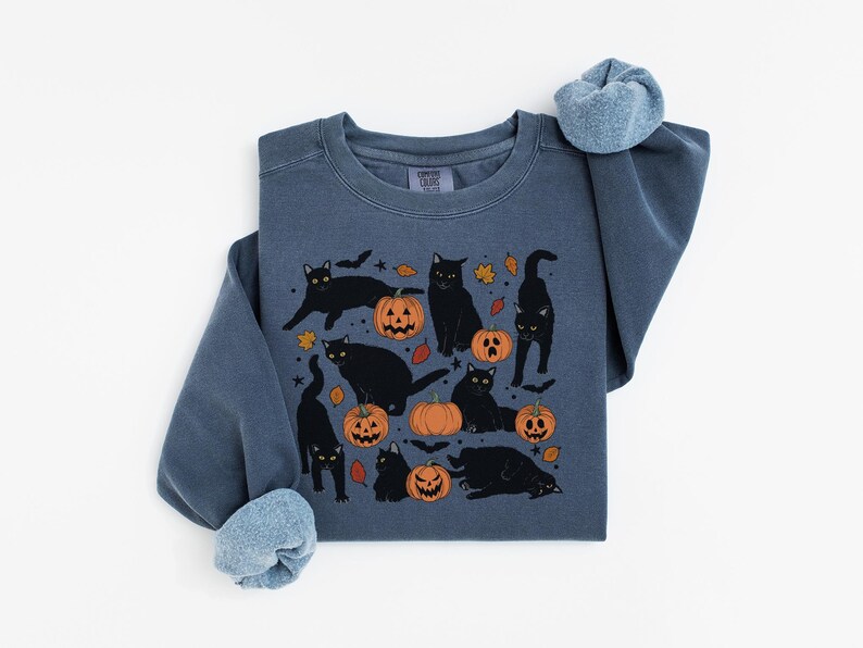 May include: A blue-grey sweatshirt featuring a Halloween design. The graphic includes black cats, orange pumpkins, and autumn leaves. The sweatshirt has a crew neck and long sleeves with rolled cuffs. The design is centered on the front of the shirt.
