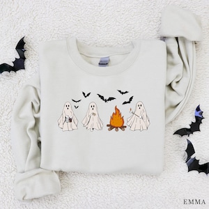 May include: A cream-coloured sweatshirt with a Halloween design. The graphic shows four cartoon ghosts gathered around a campfire, with bats flying above. The sweatshirt has long sleeves and a crew neck.