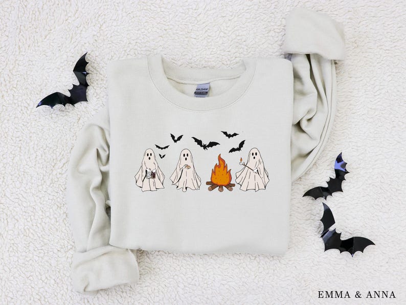 May include: A cream-coloured sweatshirt with a Halloween design. The graphic shows four cartoon ghosts gathered around a campfire, with bats flying above. The sweatshirt has long sleeves and a crew neck.