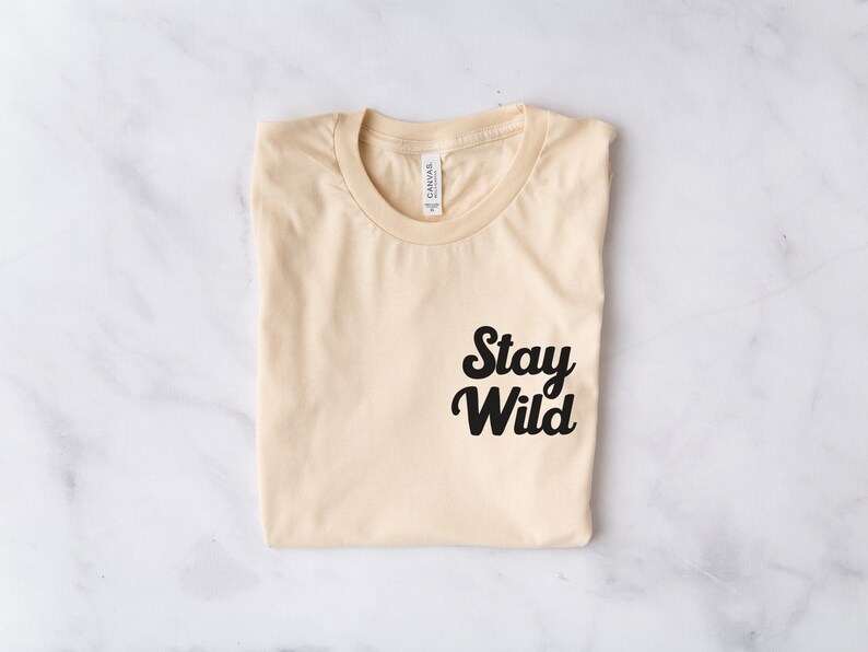 May include: A folded beige t-shirt with a black graphic that says "Stay Wild" on the front.