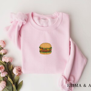 May include: A pink sweatshirt with a cartoon burger graphic on the front. The burger is yellow, brown, and green.