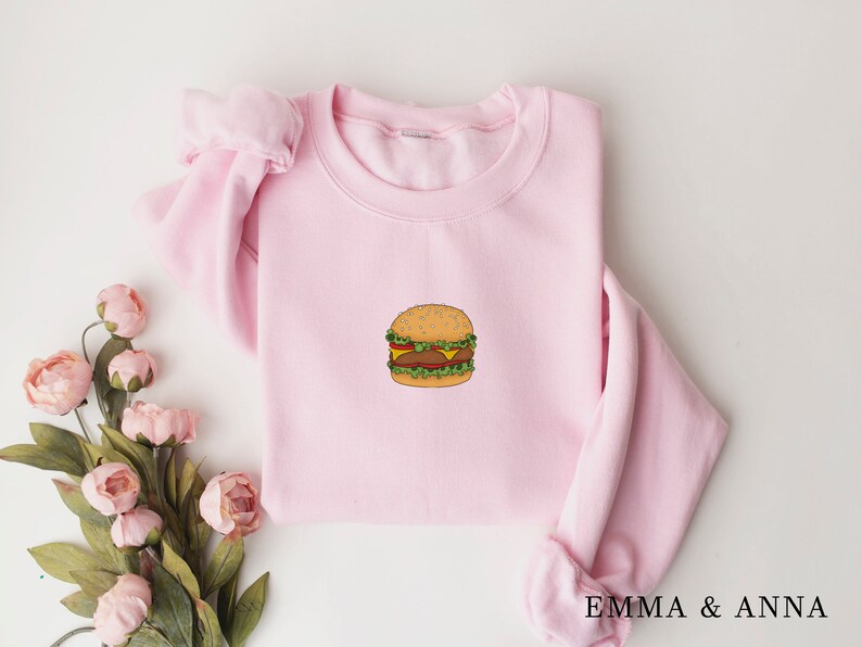 May include: A pink sweatshirt with a cartoon burger graphic on the front. The burger is yellow, brown, and green.