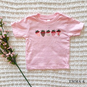 Puede incluir: Camiseta rosa de manga corta con un gr&aacute;fico de cinco fresas con rayas de diferentes colores. Las fresas tienen un coraz&oacute;n en el centro.