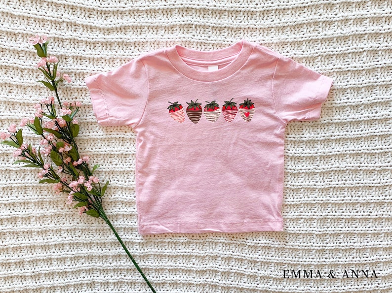 Puede incluir: Camiseta rosa de manga corta con un gr&aacute;fico de cinco fresas con rayas de diferentes colores. Las fresas tienen un coraz&oacute;n en el centro.