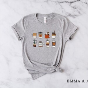 Może przedstawiać: Szary t-shirt z grafiką przedstawiającą dziewięć r&oacute;żnych napoj&oacute;w kawowych, w tym latte, kawy mrożone i latte z przyprawą dyniową.