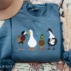 Peut inclure: Un sweat-shirt bleu avec un motif de quatre canards. Les canards sont blancs, bruns, noirs et blancs, et verts et bruns. Le sweat-shirt est pli&eacute; et pos&eacute; sur une surface &agrave; motifs.