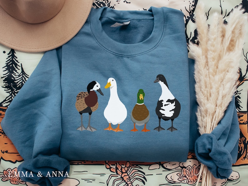 Peut inclure: Un sweat-shirt bleu avec un motif de quatre canards. Les canards sont blancs, bruns, noirs et blancs, et verts et bruns. Le sweat-shirt est pli&eacute; et pos&eacute; sur une surface &agrave; motifs.