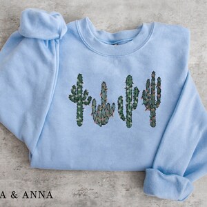 May include: Light blue sweatshirt with a graphic of four cacti with green and brown details and white lights. The cacti are arranged in a row.